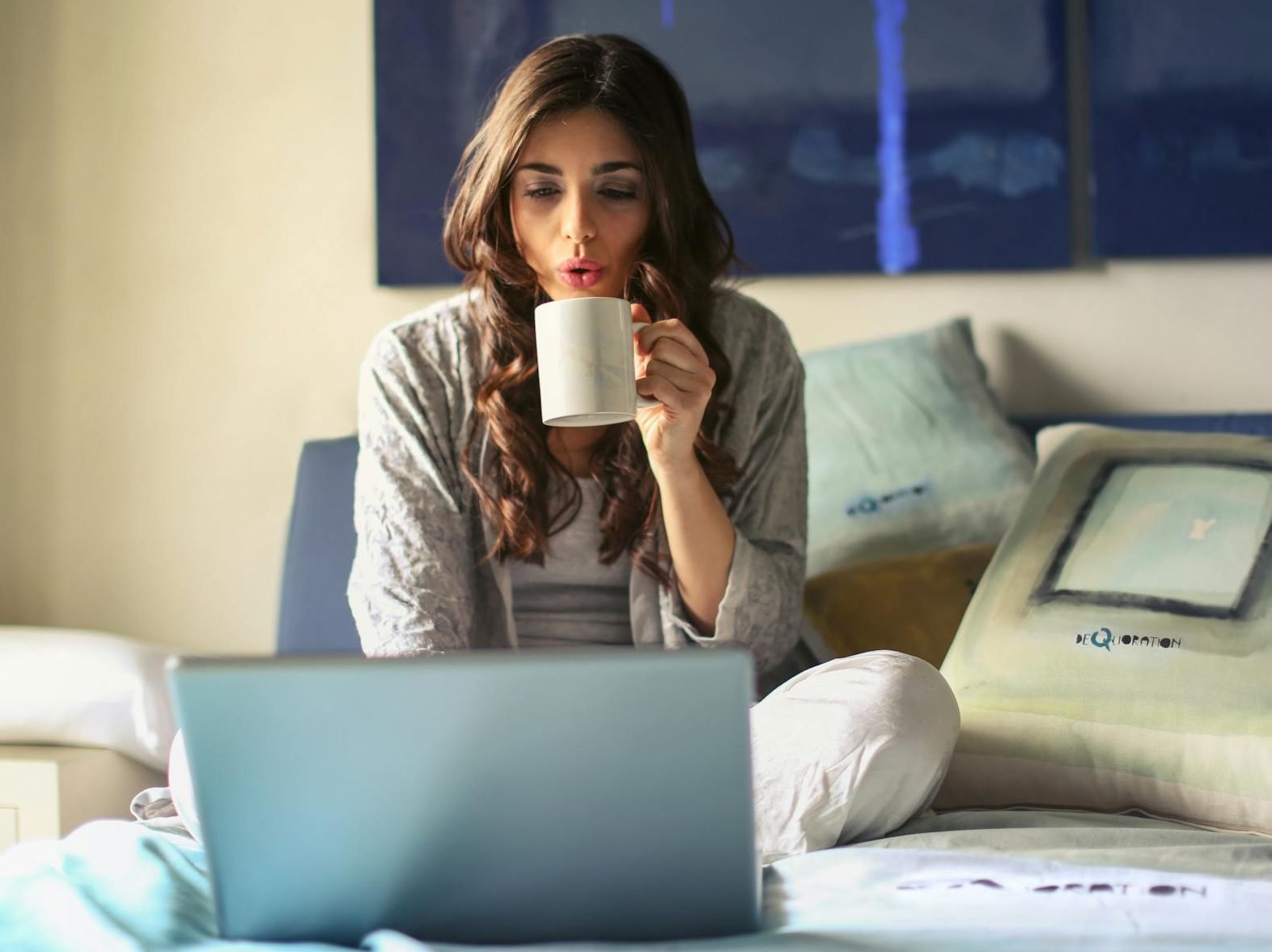 A woman enjoying coffee while working from home in a cozy bedroom setting with a laptop.